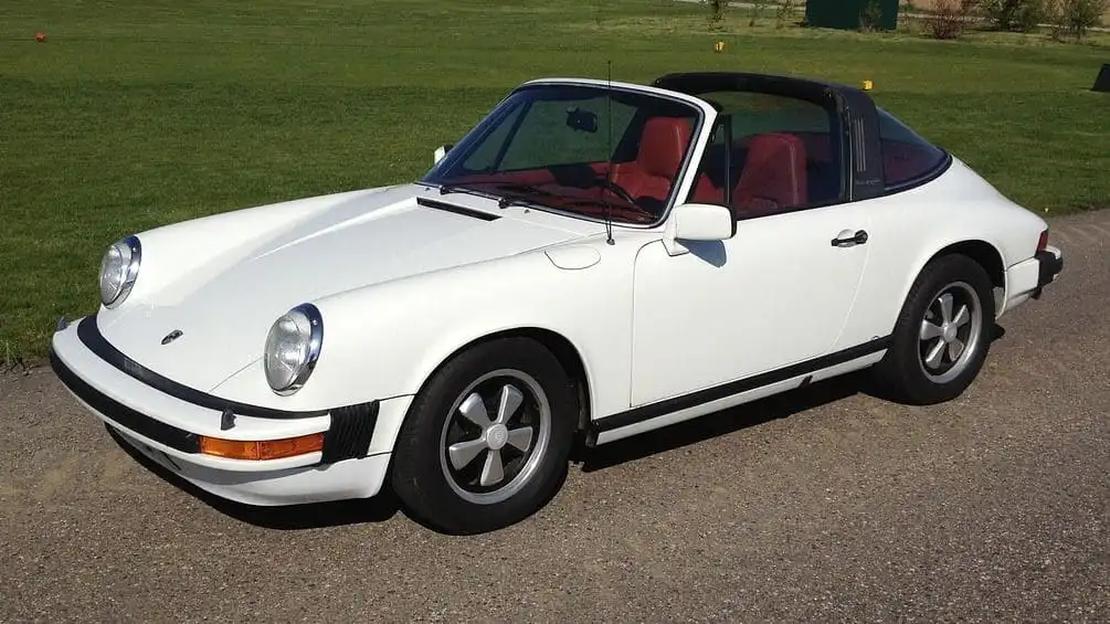 Weißer Porsche 911 Carrera 3.2 Cabriolet (1986) mit schwarzem Targa-Dach und rotem Innenraum, geparkt auf einer Landstraße.