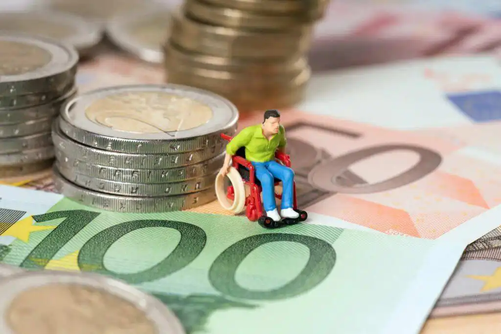 Miniaturfigur im Rollstuhl auf Euro-Banknoten und Münzen als Symbol für Krankentagegeld und finanzielle Absicherung bei Arbeitsunfähigkeit.