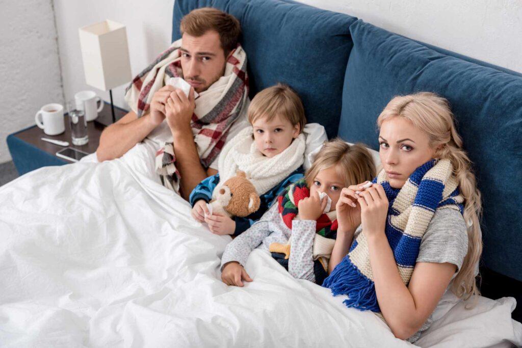 Erkrankte Familie mit Taschentüchern im Bett als Symbol für Krankenversicherung und Absicherung bei Krankheit.