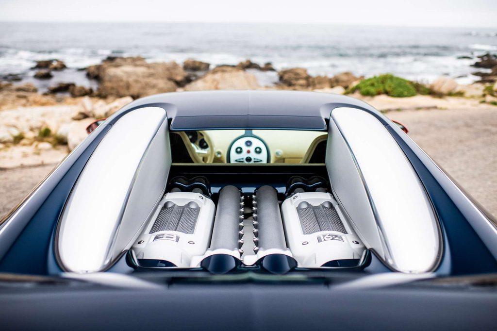 Blick auf den freiliegenden W16-Motor eines Bugatti Veyron mit sichtbaren Motorkomponenten vor einer Küstenlandschaft.